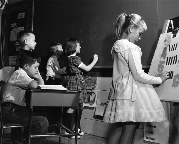 Elementary age students from the 1950s work on math problems at a chalkboard.